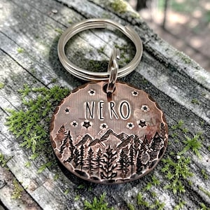 May include: A round copper-toned metal pet tag featuring a stamped mountain and pine tree forest landscape design. The name NERO is engraved in the centre above the mountains, surrounded by small star shapes. A silver metal split ring is attached.