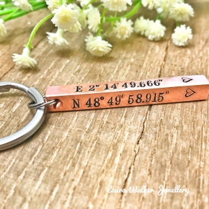 May include: A copper-coloured rectangular keyring with engraved coordinates and heart symbols. The text reads "E 2° 14' 49.666"" and "N 48° 49' 58.915"". The keyring is attached to a silver-coloured ring. The background is a wooden surface.