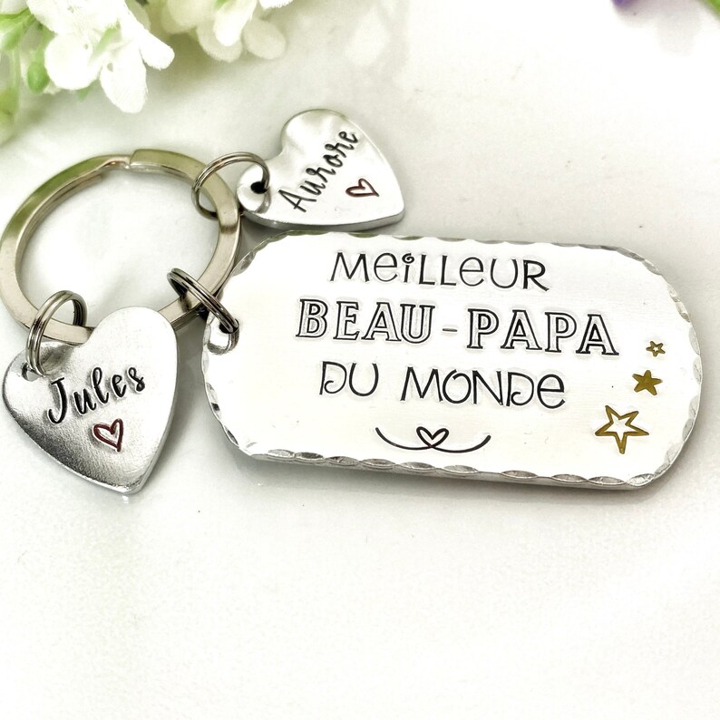 May include: A silver-toned metal keychain featuring a rectangular tag engraved with the text MEILLEUR BEAU-PAPA DU MONDE and three small stars. Two heart-shaped charms are attached to the ring, one engraved with Jules and one with Aurore.
