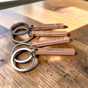 Può includere: Quattro portachiavi a barra in metallo color rame con anelli argentati. Ogni barra presenta incisioni personalizzate: le date 25.11.2019 e 16.03.2025, il testo Always S + D, numeri romani XIII.IX.MMXIX e un motivo a tacche con un cuore.