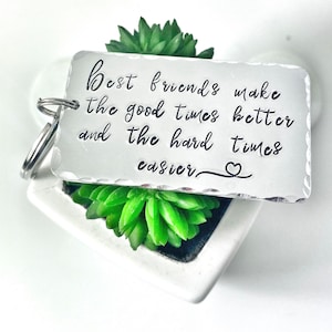 Peut inclure: Porte-clés en argent avec une citation qui dit "Les meilleurs amis rendent les bons moments meilleurs et les moments difficiles plus faciles." Le porte-clés est posé sur une surface blanche avec une petite plante succulente verte.