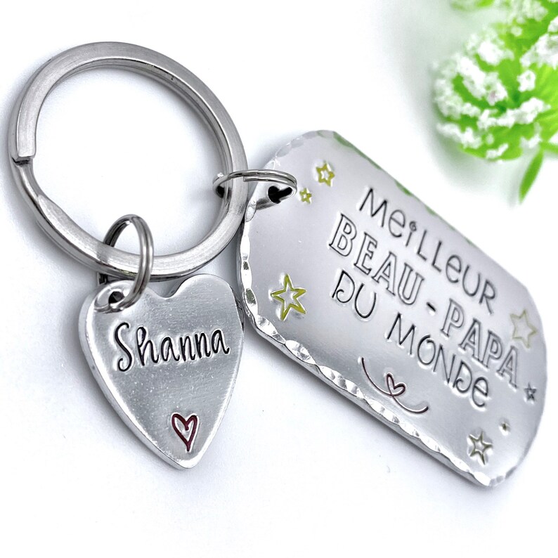 May include: A silver-toned metal keychain featuring a rectangular tag stamped with the text MEILLEUR BEAU-PAPA DU MONDE and small star icons, alongside a heart-shaped charm engraved with the name Shanna and a small red heart outline.