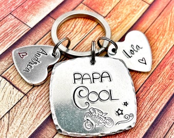 May include: A silver keychain featuring a square pendant stamped with "PAPA COOL" and a motorcycle graphic. Two heart-shaped charms are attached, one with "Andrea" and the other with "Lola".