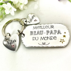 May include: A silver-toned metal keychain featuring a rectangular tag stamped with the text MEILLEUR BEAU-PAPA DU MONDE, three small stars, and a heart symbol. Two heart-shaped charms are attached, one engraved with Jules and one with Aurore.