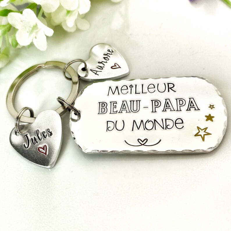 May include: A silver-toned metal keychain featuring a rectangular tag stamped with the text MEILLEUR BEAU-PAPA DU MONDE, three small stars, and a heart symbol. Two heart-shaped charms are attached, one engraved with Jules and one with Aurore.