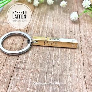 May include: A brass bar keychain with the words "LAURA" and "PAPA" engraved on it. The keychain also has a small heart symbol. The words "BARRE EN LAITON" and "BRASS BAR" are in a circle in the background.