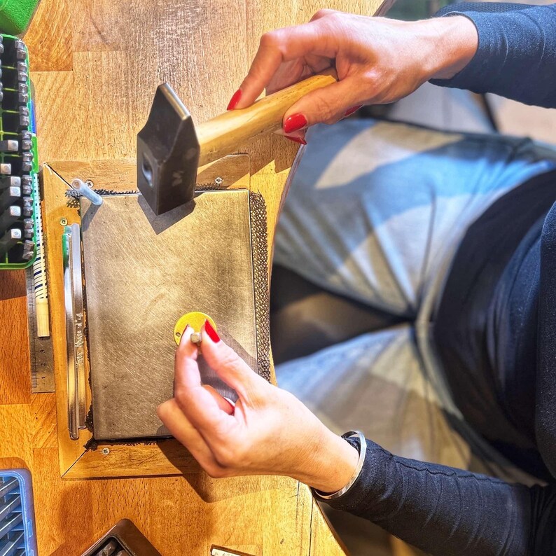 May include: A person using a hammer to shape a piece of metal on a workbench. The metal is on a gray surface with a yellow circle in the center. The person is wearing a black shirt and a bracelet.