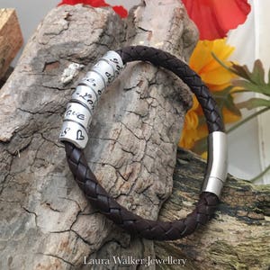 May include: A brown leather bracelet with a silver slider clasp and a series of silver beads with engraved words. The beads are arranged in a circle around the bracelet. The bracelet is lying on a piece of wood.