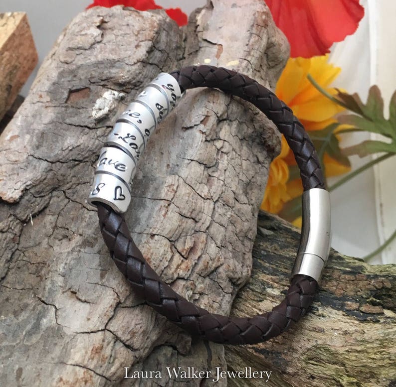 May include: A brown leather bracelet with a silver slider clasp and a series of silver beads with engraved words. The beads are arranged in a circle around the bracelet. The bracelet is lying on a piece of wood.