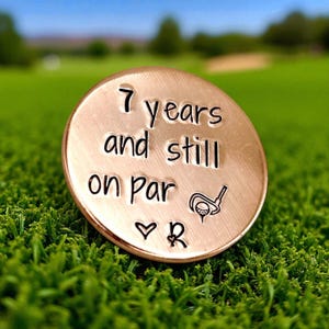 May include: A round, copper-coloured metal golf ball marker featuring the stamped text 7 years and still on par, accompanied by a small engraved golf club and ball icon, a heart symbol, and the letter R, all centred on the smooth metallic surface.