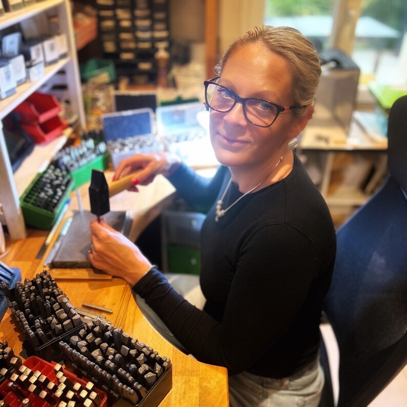 May include: A woman wearing glasses and a black shirt is working at a workbench. She is using a hammer to strike a metal stamp. The workbench is covered with various tools and supplies, including a box of metal stamps.