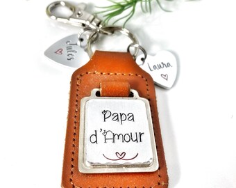 May include: A tan leather keyring featuring a rectangular metal plate engraved with the text Papa d'Amour and a small heart symbol. Two additional heart-shaped metal charms are attached, each engraved with a name, Jules and Laura, and a heart.