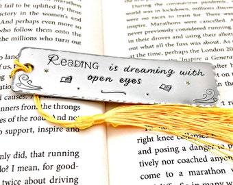 May include: A silver bookmark with a yellow tassel. The bookmark has a wavy design on the top and bottom and the text "Reading is dreaming with open eyes" in a stylized font. There are two small book icons and a star on the bookmark.