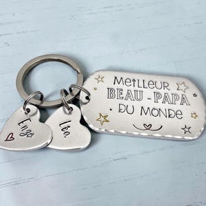 May include: A silver-toned metal keychain featuring a rectangular tag engraved with the text MEILLEUR BEAU-PAPA DU MONDE, surrounded by small star and heart symbols. Two smaller heart-shaped charms are attached, engraved with the names Enzo and L&eacute;a.