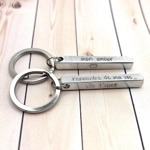 May include: Two silver-toned metal bar keychains with circular rings. One bar is engraved with "mon amour" and a heart symbol. The other bar is engraved with "rencontre de ma vie..." and "Je t'aime" in black lettering on the brushed metal surface.