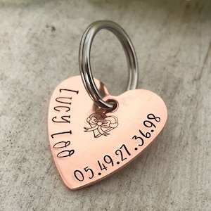 Może przedstawiać: Miedziany, sercowaty brelok do kluczy dla zwierząt z srebrnym kółkiem. Brelok jest grawerowany tekstem "Lucy Lou 05.49.21.36.98" i małym obrazkiem serca z kokardką.