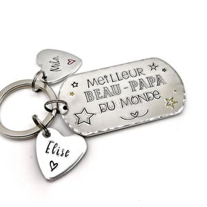 May include: A silver-toned metal keychain featuring a rectangular tag stamped with the text MEILLEUR BEAU-PAPA DU MONDE and star motifs. Two smaller heart-shaped tags are attached to the ring, each engraved with the names Mila and Elise.
