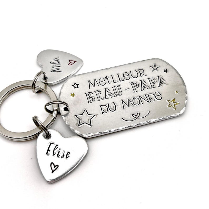 May include: A silver-toned metal keychain featuring a rectangular tag stamped with the text MEILLEUR BEAU-PAPA DU MONDE and star motifs. Two smaller heart-shaped tags are attached to the ring, each engraved with the names Mila and Elise.