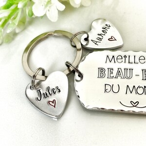 May include: A silver-toned metal keychain featuring a rectangular tag with scalloped edges and two heart-shaped charms. The rectangular tag is engraved with the text MEILLEUR BEAU-P&Egrave;RE DU MONDE. The two heart charms are engraved with the names Jules and Aurore.
