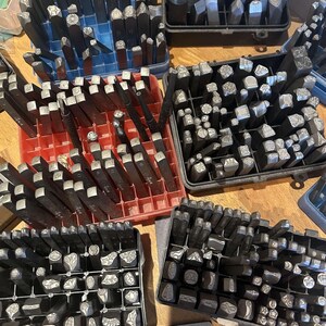 May include: Assortment of metal stamp sets in various sizes and shapes, organized in black plastic trays. The stamps are used for crafting and jewelry making.