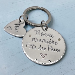 May include: A silver-toned metal keyring featuring a large circular disc with hammered edges, engraved with the text "Bonne première Fête des Pères" and two stars, plus a smaller heart-shaped charm engraved with the name "Eléna" and footprints.