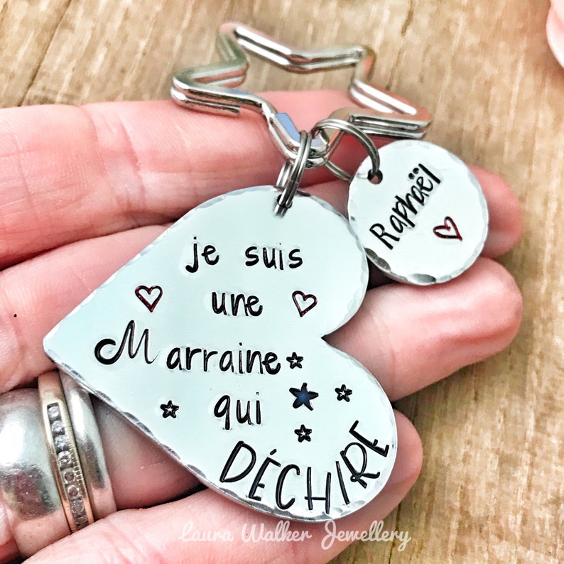 K&ouml;nnte beinhalten: Silberner herzf&ouml;rmiger Schl&uuml;sselanh&auml;nger mit dem Text "Je suis une marraine qui d&eacute;chire" und einem kleinen silbernen Kreis mit dem eingravierten Namen "Rapha&euml;l".
