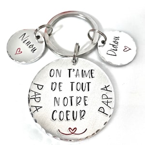 Peut inclure: Porte-clés argenté avec trois étiquettes rondes. L'étiquette centrale indique "On t'aime de tout notre coeur" avec un petit cœur en bas. Les deux autres étiquettes indiquent "Ninou" et "Didou" avec un petit cœur sur chacune.