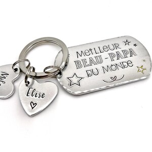 May include: A silver-toned metal keychain featuring a rectangular tag engraved with the text MEILLEUR BEAU-PAPA DU MONDE, accompanied by star motifs and two heart-shaped charms. One heart charm is engraved with the name Elise and a small heart symbol.