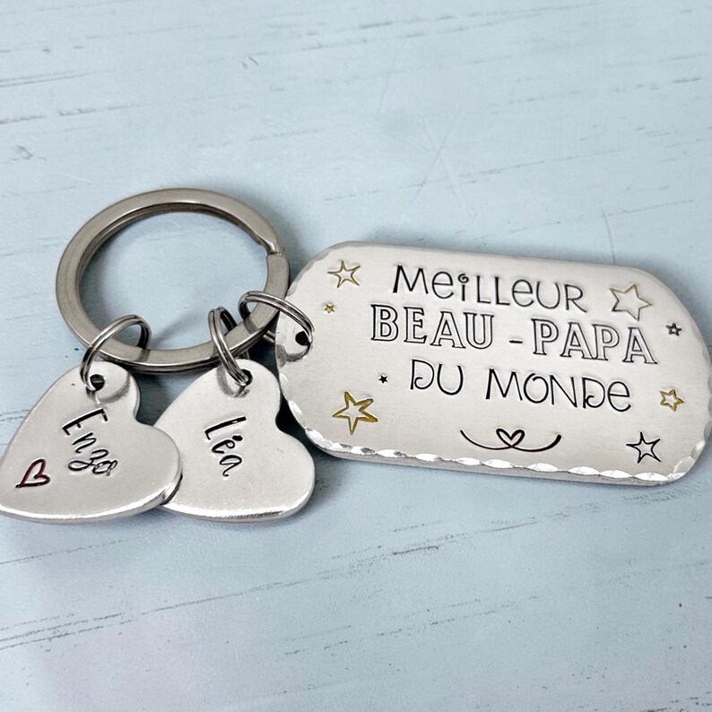 May include: A silver-toned metal keychain featuring a rectangular tag engraved with the text MEILLEUR BEAU-PAPA DU MONDE, surrounded by small star and heart symbols. Two smaller heart-shaped charms are attached, engraved with the names Enzo and L&eacute;a.