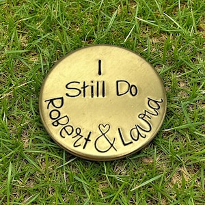 Peut inclure: Un disque rond en métal doré avec les mots "I Still Do" au centre. Les noms "Robert & Laura" sont inscrits sur le bord, séparés par un symbole de cœur. Le disque repose sur un lit d'herbe verte.