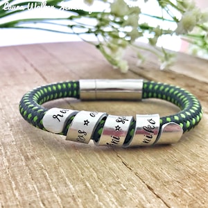 May include: A green and black braided bracelet with a silver magnetic clasp. The bracelet has four silver bands with the names "Nora", "Jess", "Lani", and "Nikko" engraved on them.