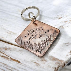 May include: A copper-coloured dog tag featuring a mountain and forest design. The tag is engraved with the word "POCKET" and has a silver key ring attached.