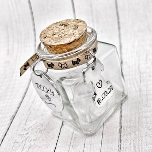 Peut inclure: Un bocal en verre transparent avec un couvercle en liège et un ruban brun avec des empreintes de pattes. Le bocal a deux étiquettes métalliques suspendues. Une étiquette dit "Rocky" avec une empreinte de patte et l'autre étiquette dit "16.09.23" avec un cœur.