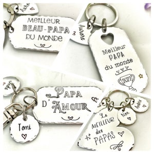 May include: A collection of silver-toned metal keychains featuring rectangular and heart-shaped tags with hand-stamped text including "MEILLEUR BEAU-PAPA DU MONDE," "Meilleur PAPA du monde," "PAPA D'AMOUR," and "Le MEILLEUR des PAPAS" with small charms.