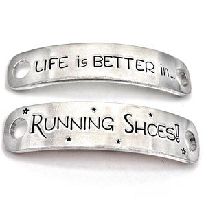 May include: Two silver metal shoe tags with the text "LIFE is BETTER in..." and "RUNNING SHOES!" in black lettering. The tags have rounded ends with holes for attaching to shoelaces. Small star accents are included.