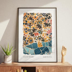 Könnte beinhalten: Gerahmter Kunstdruck mit einem Mosaikdesign in Gelb-, Blau- und Rottönen. Der Druck trägt den Text "GAUDI MOSAIC BARCELONA". Er wird auf einem Holzschrank mit einer Pflanze und einer Holzhand-Skulptur präsentiert.