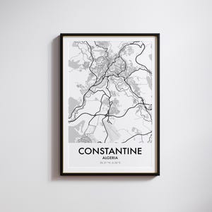 Op de afbeelding: Een ingelijste zwart-wit kaartprint van Constantine, Algerije. De kaart toont een gedetailleerde weergave van de stad, met wegen en stedelijke gebieden. De naam van de stad en de coördinaten staan onderaan afgedrukt.