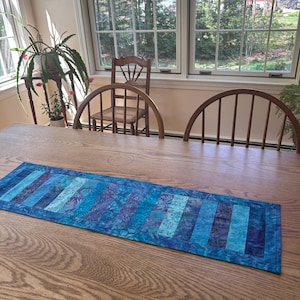 May include: A rectangular blue table runner with a patchwork design of various shades of blue and purple. The runner is placed on a wooden table, with a view of windows and chairs in the background. The runner is approximately 12 inches wide.
