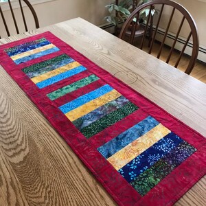 May include: A red table runner with a patchwork design featuring blue, green, and yellow fabric strips. The runner is made of batik fabric and has a quilted texture.