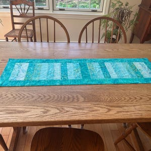 May include: A rectangular turquoise table runner with a striped pattern of light blue and green fabric. The runner is placed on a wooden table, with wooden chairs surrounding it. The room has a window in the background.