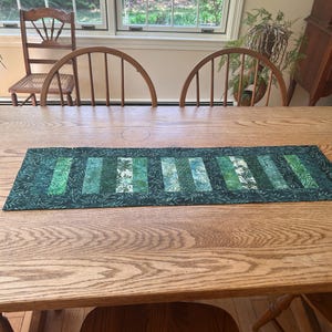 May include: A rectangular green table runner with a patchwork design of various shades of green and white floral patterns. The runner is placed on a large wooden table. Two wooden chairs are visible behind the table.