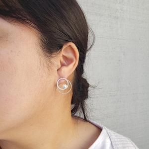 May include: Silver hoop earrings with a small pearl accent. The earrings are made of two circles, one slightly larger than the other, with a textured finish.