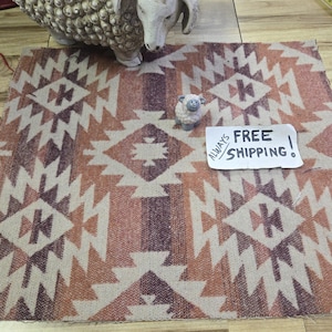 May include: A rectangular woven rug with a geometric pattern in shades of beige, orange, and purple. The rug features a repeating diamond and cross design. A small sheep figurine and a larger sheep statue are on the rug.