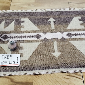 May include: A rectangular woven rug featuring a geometric pattern in shades of brown, tan, and white. The design includes repeating triangles, arrows, and chevron motifs. A small white card with the text ALWAYS FREE SHIPPING is placed on the rug.