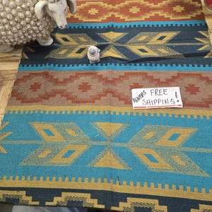 May include: A colorful woven rug with a geometric pattern in teal, brown, gold, and navy blue. The rug features diamond and cross motifs. A white sheep figurine and a small sheep figurine are on the rug.