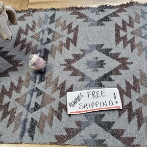 May include: A rectangular woven mat featuring a repeating geometric pattern in shades of gray, brown, and black. The mat has a fringed edge and displays a symmetrical design of diamonds and zig-zag lines across its entire surface area.