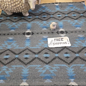 May include: Gray woven textile with a geometric pattern in black and light blue. The design features diamond and zigzag shapes. Two sheep figurines are on the textile. The text "ALWAYS FREE SHIPPING!" is on a white paper.