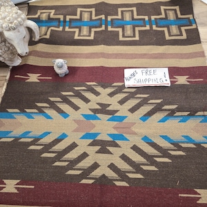 May include: A woven rug featuring a geometric pattern in brown, tan, and bright blue. The design includes repeating cross shapes and a large central diamond motif. A small white sheep figurine sits on the rug, and a white card reads ALWAYS FREE SHIPPING.
