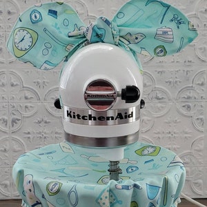 Puede incluir: Batidora de pie KitchenAid blanca con una cubierta de tela de color turquesa y blanco con estampado y un lazo de tela de color turquesa y blanco a juego en la parte superior.