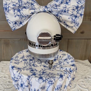 May include: A white KitchenAid stand mixer with a matching floral bow and cover. The bow and cover feature a blue floral pattern on a white background. The mixer has a silver band with the KitchenAid logo.